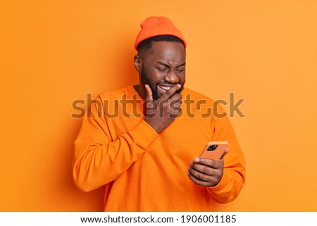 Positive dark skinned man giggles positively concentrated at smartphone screen watches funny video in internet or laughs over received message dressed in casual bright clothes isolated on orange wall Сток-фото © 