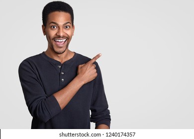 Positive dark skinned man with toothy smile, points at upper right corner with index finger, suggests use copy space, wears casual clothing, has funny expression, stands against white studio wall