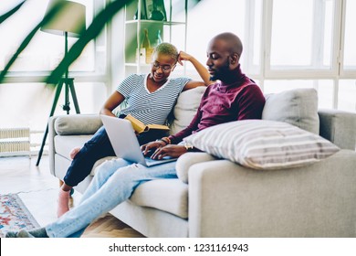 Positive dark skinned marriage spending free time at home interior, african american woman reading book while her husband typing on laptop computer blogging in social networks via wifi connection - Shutterstock ID 1231161943