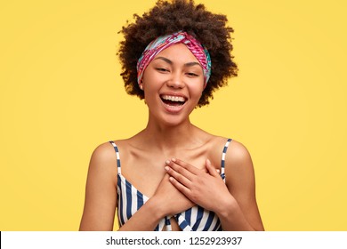 Positive dark skinned young woman with pleased expression, feels satisfied to hear compliment or confession in love, keeps both hands on chest, poses over yellow background, wears casual clothes