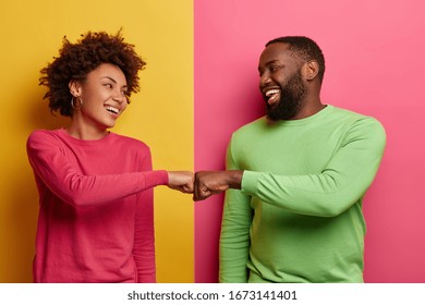 Positive dark skinned young woman and man bump fists, agree to be one team, look happily at each other, celebrates completed task, wear pink and green clothes, pose indoor, have successful deal