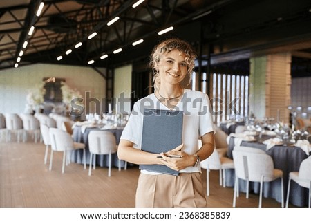 positive decorator with clipboard looking at camera near tables with festive setting in banquet hall