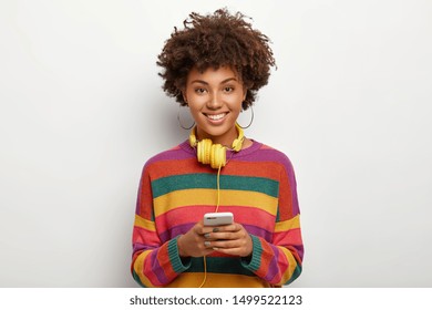 Positive delighted African American woman dressed in striped colorful sweater, holds modern cellphone connected to headphones, surfs internet, poses against white background. Technology concept