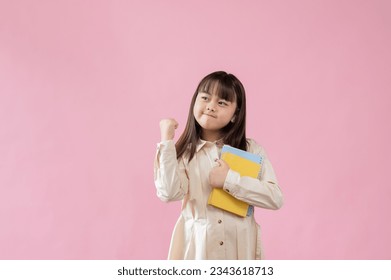 A positive and determined young Asian schoolgirl in a cute dress is holding her books and showing her clenched fist, standing against an isolated pink background. elementary school student, kids - Powered by Shutterstock