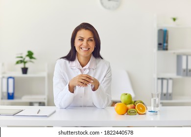 Positive Dietician Sitting At Office Desk With Fruit And Glass Of Water Looking At Web Camera And Smiling During Client's Visit, Individual Online Consultation Or While Recording Video Presentation