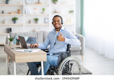 Positive disabled black man in headset using laptop computer for online work, showing thumb up gesture at home. Happy handicapped guy in wheelchair recommending remote job