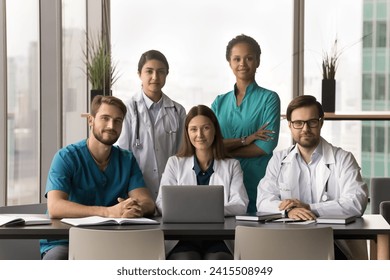 Positive diverse team of attractive young practitioners, surgeons, nurses posing together in hospital office, sitting and standing at table with laptop, looking at camera for portrait - Powered by Shutterstock