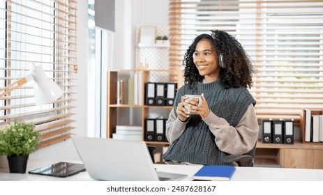 A positive, dreamy Black woman in a cozy knitted sweater, wearing headphones, is daydreaming while gazing at the view outside and having morning coffee at her desk in the office. - Powered by Shutterstock