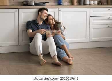 Positive dreamy married couple resting on warm floor in modern kitchen, sitting close, looking away with happy smiles, talking, discussing future plans, drinking hot beverage - Powered by Shutterstock