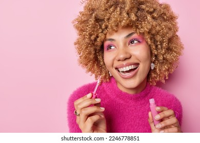 Positive dreamy woman with curly hair applies lip gloss wears vivid makeup smiles broadly has perfect white teeth wears casual pink jumper poses indoor undergoes beauty procedures wants to look nice - Powered by Shutterstock