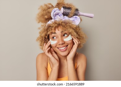 Positive dreamy young woman with curly bushy hair applies beauty patches under eyes to reduce problems of dark circles and wrinkles wears headband isolated over grey background. Skin care concept