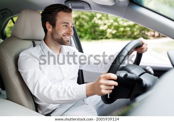 Positive driver businessman in car driving on way to\
work office, good new personal transport. Cheerful millennial\
handsome man in suit with hands on steering wheel enjoying speed\
and modern auto