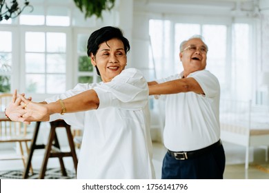 Positive Elderly Asian Man And Woman Stretching Arms And Smiling While Dancing Together In Cozy Room On Weekend Day At Home