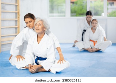 Positive elderly woman in kimono performing butterfly stretch during martial arts training. Young female coach applying pressure to knees to relieve hip tightness and improve flexibility after workout - Powered by Shutterstock