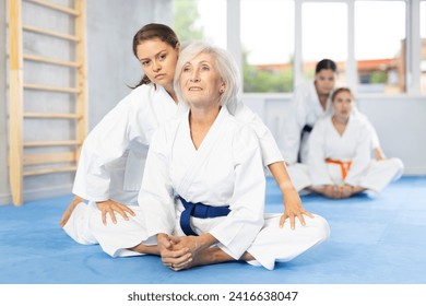 Positive elderly woman in kimono performing butterfly stretch during martial arts training. Young female coach applying pressure to knees to relieve hip tightness and improve flexibility after workout - Powered by Shutterstock