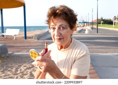 Positive elderly woman puts on make-up while looking into small powder box mirror. Beautiful woman 80 years old is resting by the sea. Concept of lifestyle, retirement time, skin aging, well being.