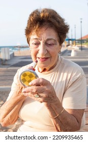 Positive elderly woman puts on lipstick while looking into small powder box mirror. Beautiful woman 80 years old is resting by the sea. Concept of lifestyle, retirement time, skin aging, well being.