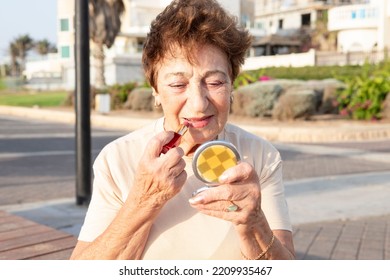 Positive elderly woman puts on lipstick while looking into small powder box mirror. Beautiful woman 80 years old is resting by the sea. Concept of lifestyle, retirement time, skin aging, well being.
