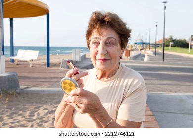 Positive elderly woman puts on make-up while looking into small powder box mirror. Beautiful woman 80 years old is resting by the sea. Concept of lifestyle, retirement time, skin aging, well being.