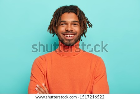 Positive emotions concept. Handsome dark skinned hipster with dreads has pleasant smile, has white teeth, happy to hear good news, wears bright clothes, isolated over blue background, has lucky day