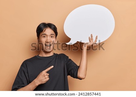 Positive emotions. Indoor photo of young glad smiling Asian man standing on left on beige background wearing casual clothes holding and pointing at white speech bubble with space for promotion