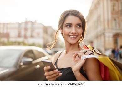 Positive emotions. Lifestyle concept. Close up of young charming dark-haired caucasian woman in black dress smiling with teeth, looking aside with relaxed expression, chatting with boyfriend on phone