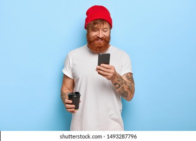 Positive emotions and modern technologies concept. Cheerful stylish man wears red hat and white t shirt, has ginger beard chats with friends via cell phone connected to wireless internet drinks coffee - Powered by Shutterstock