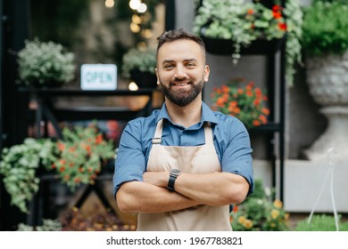 Positive Emotions Of Owner Of Studio, Cafe, Store. Cheerful Confident Millennial Handsome Bearded Man In Apron With Crossed Arms On Chest, On Flower Shop Outdoor Background, With Plants, Copy Space