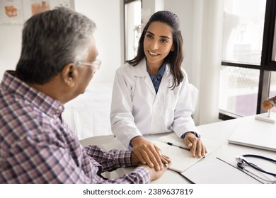 Positive empathetic young doctor woman giving support to elderly patient man with geriatric diseases, touching hand over workplace table, holding arm, expressing medical care, smiling - Powered by Shutterstock