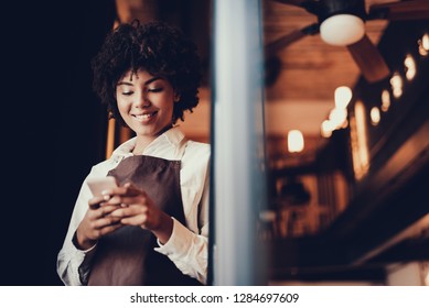 Positive employee woman holding cellular in hands and standing inside cafe. She typing message and smiling