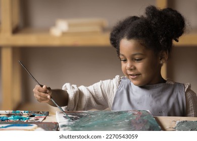 Positive engaged art school African pupil girl in apron enjoying painting on canvas, stroking with paintbrush, enjoying creativity, inspiration, training creative talent, skills - Powered by Shutterstock