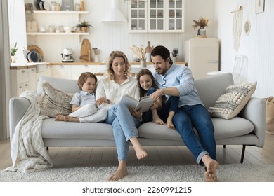 Positive engaged couple of parents teaching two cute kids to read book, watching pictures, telling funny fairytale, relaxing on home couch, hugging children, enjoying literature hobby - Powered by Shutterstock