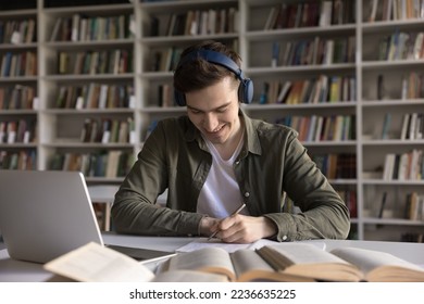 Positive engaged student guy in wireless headphones listening online learning course, webinar at laptop, writing notes, summary, essay, article, studying at table with books