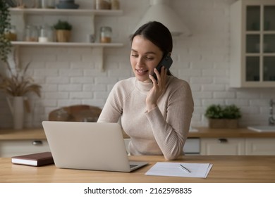 Positive Engaged Young Freelance Professional Woman Working In Home Kitchen, Making Mobile Phone Call, Speaking To Client, Using Online App On Laptop Computer, Giving Consultation, Help, Assist