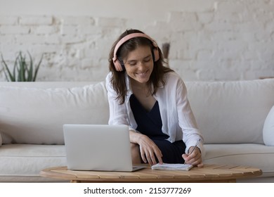 Positive Engaged Young Student Girl In Trendy Wireless Headphones Watching Online Lesson On Laptop, Listening To Audio Training, Learning Book, Writing Notes, Preparing For College Test