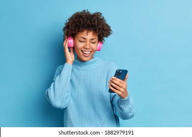 Positive entertained millennial girl with curly hair holds smartphone makes video call wears stereo headphones for listening favorite music dressed in warm jumper isolated over blue background - Shutterstock ID 1882979191