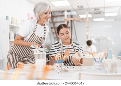 Positive enthusiastic senior woman offering guidance to interested young girl painting ceramic mug in pottery class. Creative hobby concept.. - Powered by Shutterstock