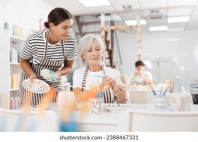 Positive enthusiastic young girl assisting interested senior woman painting ceramic mugs and plates in pottery class. Creative hobby concept.. - Powered by Shutterstock