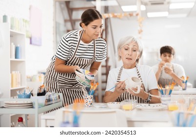 Positive enthusiastic young girl assisting interested senior woman painting ceramic mugs and plates in pottery class. Creative hobby concept.. - Powered by Shutterstock