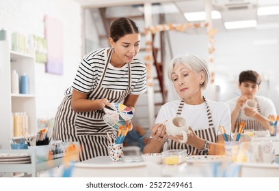 Positive enthusiastic young girl assisting interested senior woman painting ceramic mugs and plates in pottery class. Creative hobby concept.. - Powered by Shutterstock