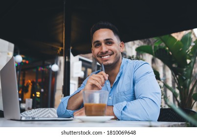 Positive Ethnic Male Freelancer In Casual Wear Smiling And Looking At Camera While Working On Remote Project On Laptop In Cafe