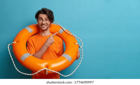 Positive European man lifesaver poses with inflated orange life ring, indicates at empty space, shows way to sea or swimming pool. Lifeguard on duty holds rescue circle. Safety beach holiday.