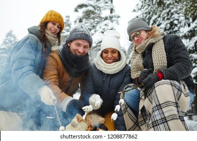 Positive Excited Young Multi-ethnic Friends In Winter Coats Sitting By Campfire And Roasting Marshmallows On Fire At Camping In Forest