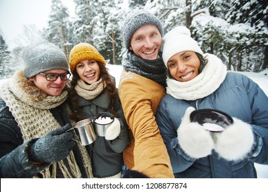 Positive Excited Young Multi-ethnic Friends In Warm Clothing Holding Thermos Mugs And Taking Selfie Together In Winter Forest, They Hiking Together