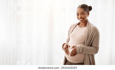 Positive expecting millennial african american woman hold hands in heart shape on her big tummy, smiling at camera. Happy young black pregnant lady posing next to window at home, banner, copy space - Powered by Shutterstock