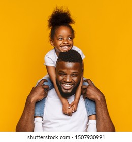 Positive family. Cheerful african dad riding his cute little daughter on shoulders, orange studio background