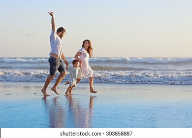 Positive family - father, mother with baby son hold hands and run with fun along edge of sea on smooth sand beach. Active parents and people outdoor activity on tropical summer holidays with children