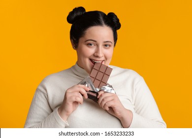 Positive Fatty Girl Biting Chocolate Bar, Enjoying Eating Sweets, Posing Over Yellow Background And Looking At Camera, Free Space
