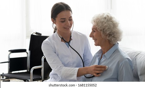 Positive female caregiver visit smiling senior disabled grandmother check heart rate, young woman doctor use statoscope do regular checkup of old lady patient at home, elderly healthcare concept