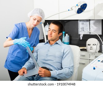 Positive female cosmetologist giving consultation to young man patient lying on couch in beautician office of aesthetic clinic